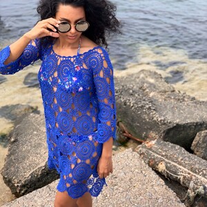 K&ouml;nnte beinhalten: Eine Frau tr&auml;gt ein blaues H&auml;kelkleid mit einem runden, offenen Muster. Sie steht auf einer Betonfl&auml;che in der N&auml;he des Wassers.