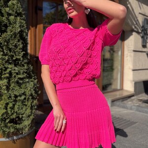 May include: A vibrant fuchsia two-piece knit set. The top features a crew neck and short sleeves with a textured, openwork design. The matching skirt is a mini length with a pleated hem. The outfit is a bright, eye-catching ensemble.
