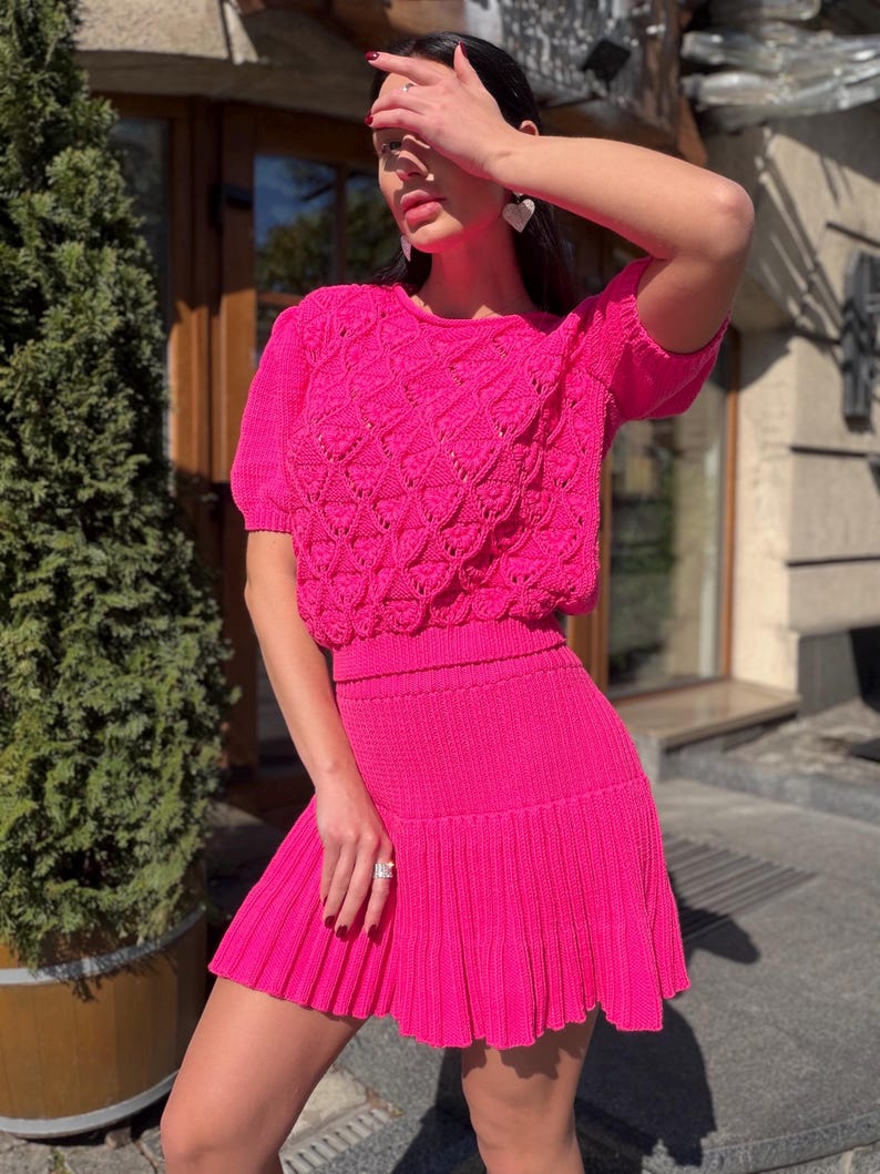 May include: A vibrant fuchsia two-piece knit set. The top features a crew neck and short sleeves with a textured, openwork design. The matching skirt is a mini length with a pleated hem. The outfit is a bright, eye-catching ensemble.