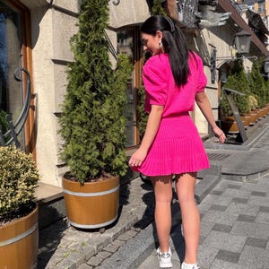 May include: A bright pink, two-piece outfit consisting of a short-sleeved top and a pleated mini skirt. The outfit is worn with white sneakers. The background features a building with a stone facade and potted plants.