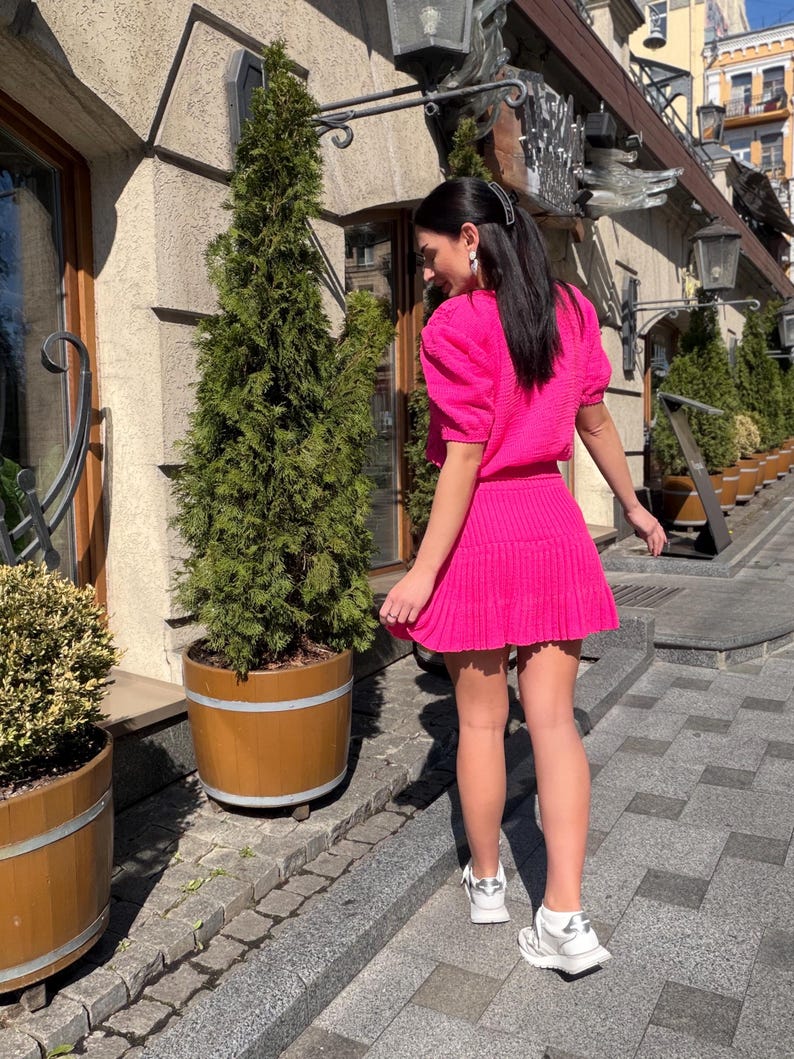 May include: A bright pink, two-piece outfit consisting of a short-sleeved top and a pleated mini skirt. The outfit is worn with white sneakers. The background features a building with a stone facade and potted plants.
