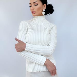 May include: A white ribbed turtleneck sweater with long sleeves. The sweater is fitted and has a high neckline.
