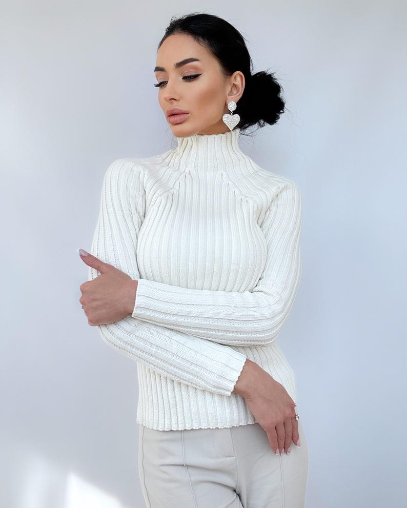 May include: A white ribbed turtleneck sweater with long sleeves. The sweater is fitted and has a high neckline.