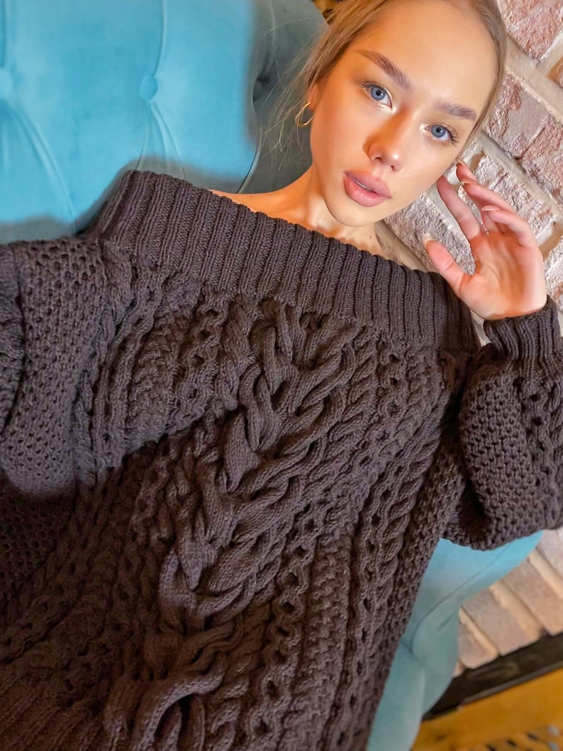 May include: A dark brown long-sleeve sweater featuring a wide ribbed neckline and a prominent vertical cable knit pattern down the center. The garment is constructed with a thick, textured knit fabric and includes ribbed cuffs at the wrists.