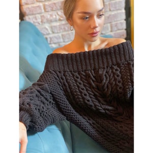 May include: A dark charcoal gray off-the-shoulder sweater featuring a chunky knit texture with vertical cable knit patterns and a wide ribbed neckline. The garment has long sleeves with ribbed cuffs and a thick, textured weave throughout the body.