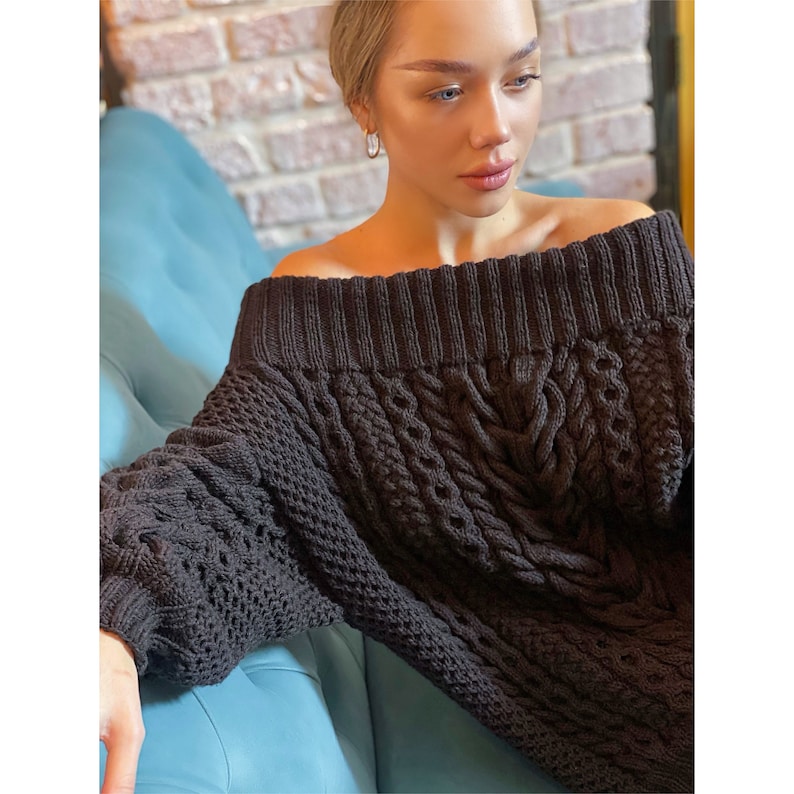 May include: A dark charcoal gray off-the-shoulder sweater featuring a chunky knit texture with vertical cable knit patterns and a wide ribbed neckline. The garment has long sleeves with ribbed cuffs and a thick, textured weave throughout the body.