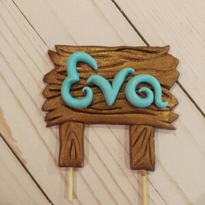 Puede incluir: Un letrero de madera marr&oacute;n con el nombre "Eva" en azul claro, montado sobre dos palitos de madera. El letrero tiene un aspecto de veta de madera texturizada, y las letras est&aacute;n en una fuente cursiva.