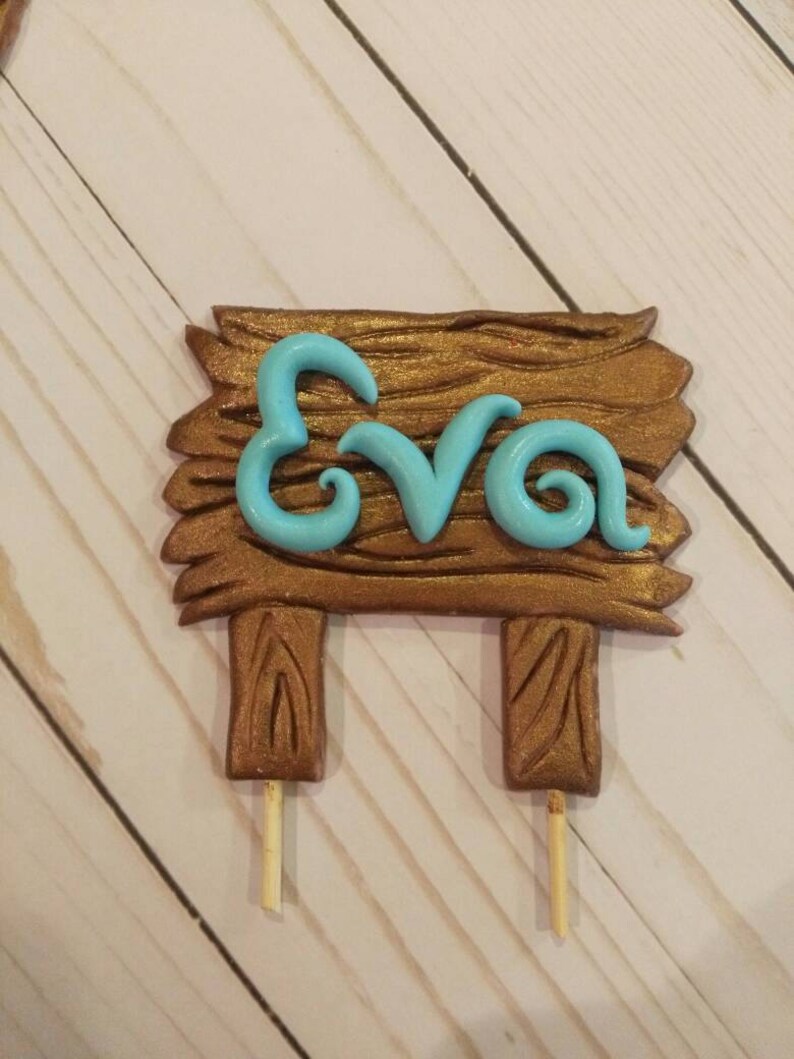 Puede incluir: Un letrero de madera marr&oacute;n con el nombre "Eva" en azul claro, montado sobre dos palitos de madera. El letrero tiene un aspecto de veta de madera texturizada, y las letras est&aacute;n en una fuente cursiva.