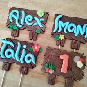 Puede incluir: Cuatro letreros de madera marr&oacute;n con texto azul, celeste y naranja y detalles decorativos. Los letreros dicen "Alex", "Mona", "Talia" y "1". Cada letrero tiene un palo de madera adjunto en la parte inferior.