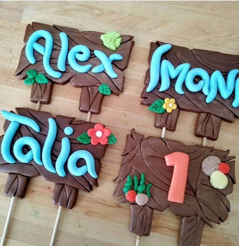 Puede incluir: Cuatro letreros de madera marr&oacute;n con texto azul, celeste y naranja y detalles decorativos. Los letreros dicen "Alex", "Mona", "Talia" y "1". Cada letrero tiene un palo de madera adjunto en la parte inferior.