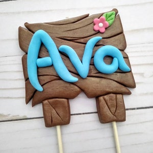 Puede incluir: Un letrero de madera marr&oacute;n con el nombre "Ava" en azul claro, coronado con una flor rosa y una hoja verde. El letrero est&aacute; sostenido por dos palitos de madera, ideal para un adorno de pastel.