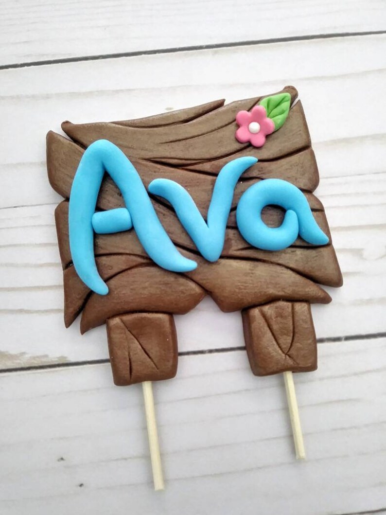 Puede incluir: Un letrero de madera marr&oacute;n con el nombre "Ava" en azul claro, coronado con una flor rosa y una hoja verde. El letrero est&aacute; sostenido por dos palitos de madera, ideal para un adorno de pastel.