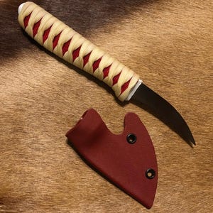 May include: A curved, black blade knife with a cream-colored handle wrapped in red cord. The knife comes with a red leather sheath.