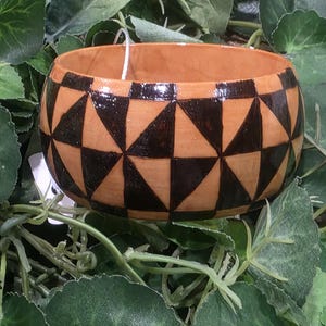 May include: A round, handcrafted bowl with a geometric design. The bowl features a natural wood tone base with a repeating pattern of dark brown triangles and squares around the rim. The bowl is surrounded by green leaves.
