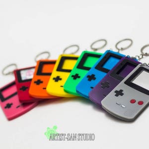 May include: A collection of colorful Game Boy keychains in pink, red, orange, yellow, green, blue, purple, and gray. Each keychain features a silver keyring and a miniature replica of the classic handheld console. The keychains are arranged on a white surface.
