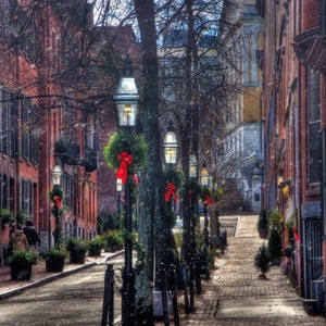 May include: A snowy street lined with brick buildings and decorated with red and green Christmas wreaths.  The street is illuminated by streetlights with a vintage design.