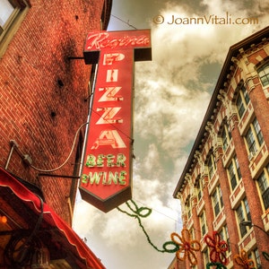 May include: A red and white neon sign with the words "Regina Pizza Beer & Wine" on it. The sign is mounted on the side of a brick building. The building has a red awning and a window with white blinds.
