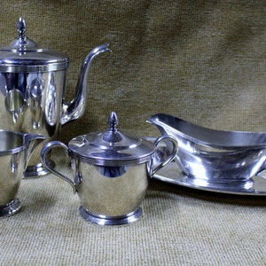 May include: A silver tea set including a teapot, creamer, sugar bowl, and gravy boat with a matching oval tray. The set is made of shiny metal and sits on a textured surface.