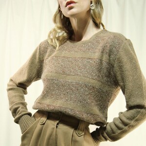 May include: A tan sweater with horizontal stripes of mixed colours, worn with high-waisted, pleated tan trousers. The sweater has long sleeves and a round neckline. The trousers have button details.