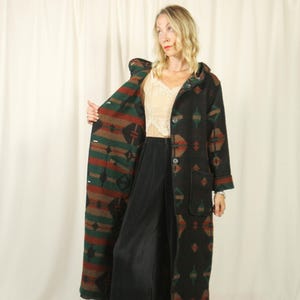 May include: A long, black, hooded coat featuring a geometric pattern in shades of dark green and rust red. The coat has a button-front closure, long sleeves, and two large patch pockets on the front. It is worn over a beige lace camisole and black trousers.