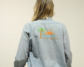 May include: A light blue button-down shirt with embroidered details on the back. The embroidery features a palm tree, a setting sun, and ocean waves. There are also smaller embroidered details of a snail and a butterfly on the shirt.