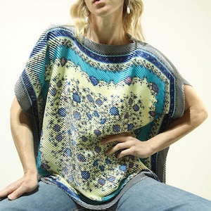 May include: A patterned, pleated top in yellow, blue, and grey. The top has a floral design and a round neckline. The model is wearing blue jeans.