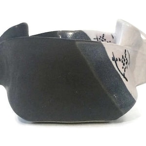 May include: A black and white ceramic bowl with a unique, asymmetrical design. The bowl has a textured surface and a slightly raised rim.