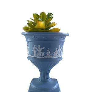 Op de afbeelding: Een lichtblauwe keramische plantenpot met een vierkante basis en een wit ontwerp van figuren en een vuur. De pot is gevuld met een groene succulent.