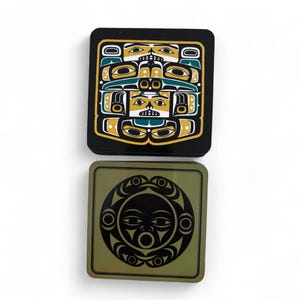 Puede incluir: Dos posavasos cuadrados con dise&ntilde;os de inspiraci&oacute;n nativa americana. Uno es negro con un dise&ntilde;o amarillo, verde azulado y blanco. El otro es verde oliva con un dise&ntilde;o de cara de sol negra. Ambos son cuadrados y brillantes.