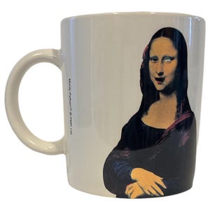 Könnte beinhalten: Weiße Keramiktasse mit einem bunten Bild der Mona Lisa mit einem überraschten Ausdruck. Die Tasse hat einen schwarzen Henkel und den Text "Monty Python © Python (UK) Ltd." auf der Seite.