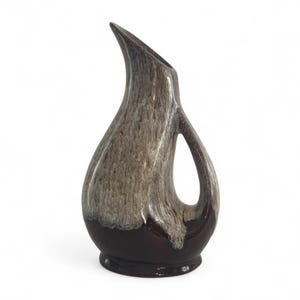 May include: A brown and tan ceramic jug with a distinctive, curved spout and a handle. The base is a darker brown, and the body has a textured, speckled appearance. The jug is designed with a modern, artistic flair.
