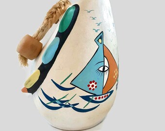 May include: White ceramic hanging ornament with a rope handle. The ornament is painted with a blue, orange, and green sailboat on a white background with blue waves and white birds.