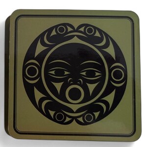 Puede incluir: Posavasos cuadrado con un dise&ntilde;o de sol negro sobre un fondo verde oliva. El sol presenta una cara con ojos, boca y elementos decorativos. El posavasos tiene un borde oscuro y probablemente est&aacute; hecho de un material duradero.