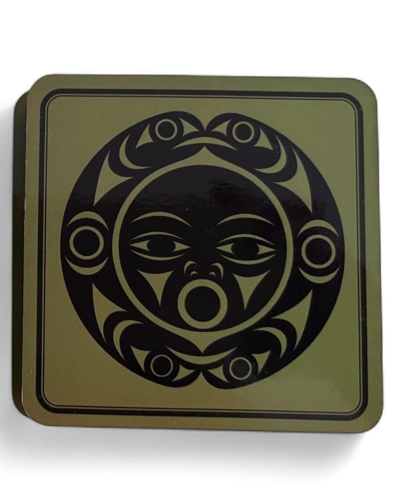 Puede incluir: Posavasos cuadrado con un dise&ntilde;o de sol negro sobre un fondo verde oliva. El sol presenta una cara con ojos, boca y elementos decorativos. El posavasos tiene un borde oscuro y probablemente est&aacute; hecho de un material duradero.