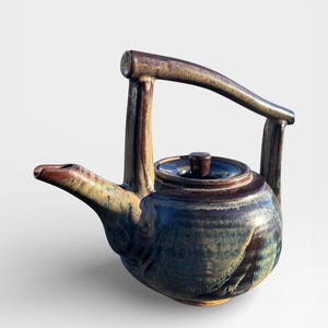 May include: A handcrafted ceramic teapot with a unique handle design. The teapot features a dark blue, brown, and tan glaze. The spout is angled, and the lid has a small, round knob. The handle is a curved, wooden-like design.