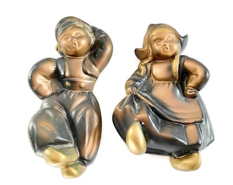 May include: A pair of vintage ceramic wall decor figurines of a boy and girl in traditional Dutch clothing. The boy is wearing a black and brown outfit with a gold hat, and the girl is wearing a black and gold dress with a white bonnet.