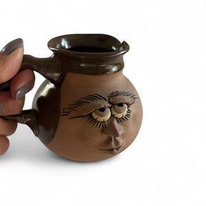 Könnte beinhalten: Eine handgefertigte braune Keramiktasse mit einem Gesicht auf der Vorderseite. Die Tasse hat einen Henkel und einen dunkelbraunen Rand. Das Gesicht hat detaillierte Augenbrauen, Augen und eine Nase. Die Tasse wird von einer Hand gehalten.