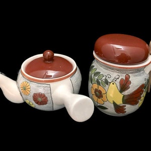 Puede incluir: Una tetera y un azucarero de cerámica blanca con tapas marrones y diseños florales y de pájaros. La tetera tiene un asa en el lateral y un pico en la parte delantera. El azucarero tiene una cuchara de madera unida a la tapa.