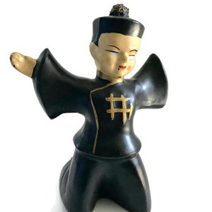 May include: A black ceramic figurine of a person wearing a black robe with gold accents. The person is kneeling with their arms outstretched. The figurine has a painted face with black hair and a gold topknot.