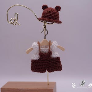 May include: A miniature crochet outfit, including a brown bear hat and a two-piece set, hangs on a small wooden hanger. The outfit is brown and white, and the hat has small bear ears. The outfit is displayed on a gold-colored stand.