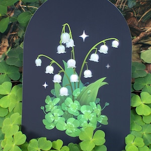 Puede incluir: Una ilustración digital de flores blancas de lirio de los valles con hojas verdes sobre un fondo negro. Las flores están rodeadas de tréboles de cuatro hojas.