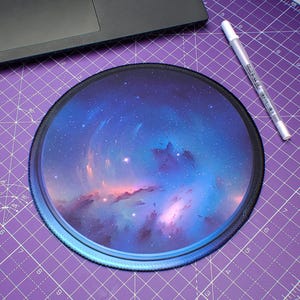 May include: Circular mouse mat with a vivid nebula design in blue, purple, and pink hues. It has a black stitched border. A white pen lies on a purple cutting mat beside the mouse mat.
