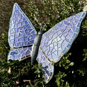 May include: A purple mosaic butterfly garden decoration with iridescent glass tiles. The butterfly is perched on a branch with green leaves.