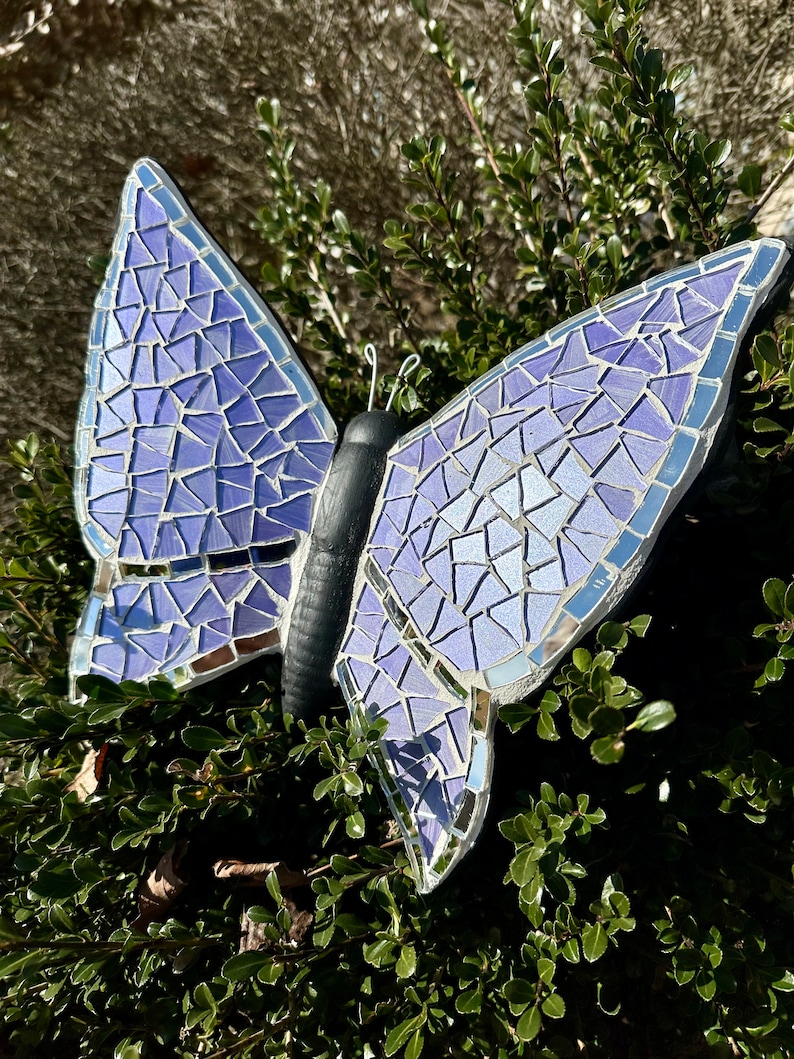 May include: A purple mosaic butterfly garden decoration with iridescent glass tiles. The butterfly is perched on a branch with green leaves.