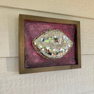 May include: A framed mosaic art piece featuring a pair of lips. The lips are made of small, reflective mirror tiles, set against a textured, deep red background. The frame is a dark, natural wood.