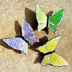 May include: Three mosaic butterfly wall decorations. The butterflies are made of small, iridescent tiles in shades of purple, green, and yellow. The butterflies are arranged in a triangular pattern on a light brown background.