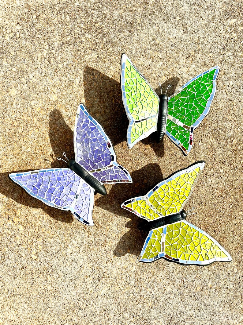 May include: Three mosaic butterfly wall decorations. The butterflies are made of small, iridescent tiles in shades of purple, green, and yellow. The butterflies are arranged in a triangular pattern on a light brown background.