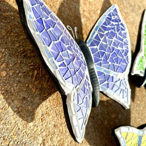 May include: A purple mosaic butterfly with black accents and silver detailing. The butterfly is made of small, iridescent tiles and has a textured surface.