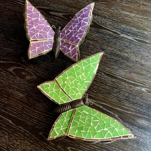 May include: Two mosaic butterflies, one purple and one green, on a dark wood background.