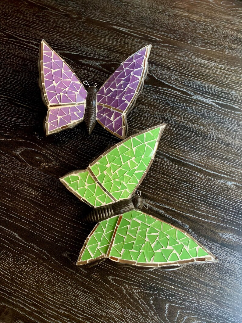 May include: Two mosaic butterflies, one purple and one green, on a dark wood background.